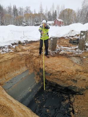 Kuvassa kaivanto, jonka pohjalla mustaa sulfidista sedimenttiä ja tutkija mittaamassa kaivannon syvyyttäkav