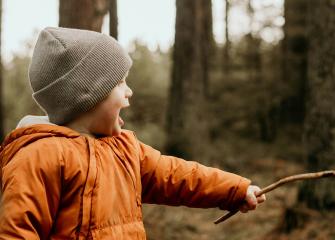 Pieni lapsi pitää kiinni kepistä ja nauraa metsässä.