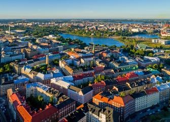 Ilmakuva tiiveistä kaupunkikortteleista, punakattoisista ja vaaleista kerrostaloista. Taustalla näkyy vesialuetta ja puistoja.