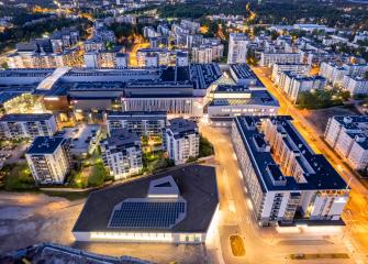 Ilmakuva tiiviistä kaupunkialueesta iltahämärässä: kerrostaloja, valaistuja katuja ja suuri keskusrakennus, jonka etualan katolla näkyy aurinkopaneeleja.