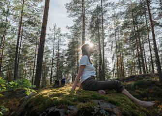 Henkilö istuu paljain jaloin sammaleisella kalliolla aurinkoisessa mäntymetsässä. Auringonvalo siivilöityy puiden välistä.