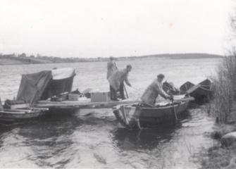 Hydrological measurements on the River Kemijoki in 1956. Several wooden boats are moored at the riverbank and connected by a makeshift working platform. Three people are carrying out field measurements, with a tent and measuring equipment visible on the platform. The black-and-white photograph documents hydrological fieldwork on a windy river.