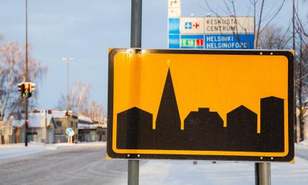 Valokuvassa on etualalla taajama-liikennemerkki ja taustalla näkyy talvista maisemaa sekä kaistaopasteet, joissa lukee Keskusta ja Helsinki.