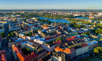 Ilmakuva tiiveistä kaupunkikortteleista, punakattoisista ja vaaleista kerrostaloista. Taustalla näkyy vesialuetta ja puistoja.