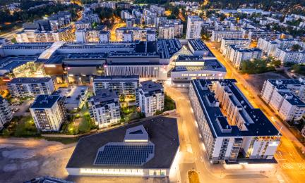 Ilmakuva tiiviistä kaupunkialueesta iltahämärässä: kerrostaloja, valaistuja katuja ja suuri keskusrakennus, jonka etualan katolla näkyy aurinkopaneeleja.