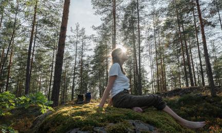Henkilö istuu paljain jaloin sammaleisella kalliolla aurinkoisessa mäntymetsässä. Auringonvalo siivilöityy puiden välistä.