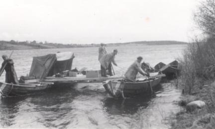 Hydrological measurements on the River Kemijoki in 1956. Several wooden boats are moored at the riverbank and connected by a makeshift working platform. Three people are carrying out field measurements, with a tent and measuring equipment visible on the platform. The black-and-white photograph documents hydrological fieldwork on a windy river.