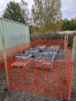 Kuvassa harmaita kannellisia muovilaatikoita oranssin huomioaidan takana